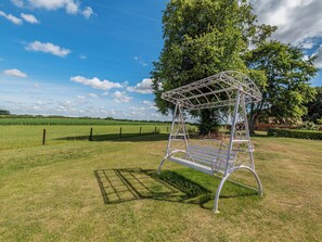 A white metal swing chair in a grassy field with a tree and a fence in the background.