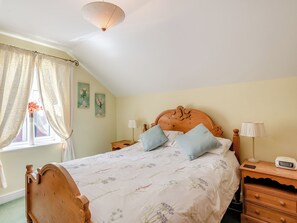 Een slaapkamer met een houten hoofdbord, bloemenbedrukte beddengoed, twee bedlampjes, een nachtkastje met een lamp en een raam met gordijnen.
