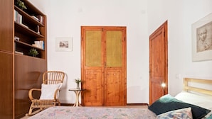 Un dormitorio con cama, un armario de madera, una silla de mimbre, una mesita pequeña y un par de puertas.