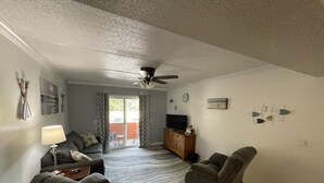 Uma sala de estar com ventilador de teto, um sofá, duas poltronas, uma TV e uma porta de vidro deslizante que dá para uma área externa.