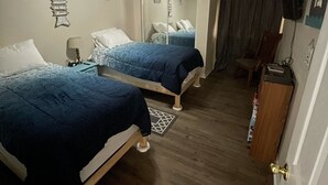 Quarto de hotel com duas camas, um ventilador de teto, espelho, televisão e piso de madeira.
