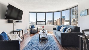 A modern living room with a large TV, a grey sofa, a coffee table, and a view of the city through large windows.