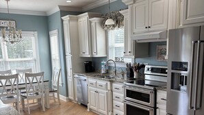 A modern kitchen with white cabinetry, granite countertops, stainless steel appliances, and a dining area with white chairs. - Custom Home on LakeFront in Ocean Lakes (Myrtle Beach)