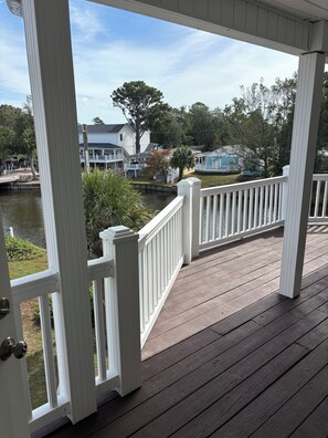 A wooden deck with white railings overlooking a calm body of water, with houses and trees in the background. - Custom Home on LakeFront in Ocean Lakes (Myrtle Beach)