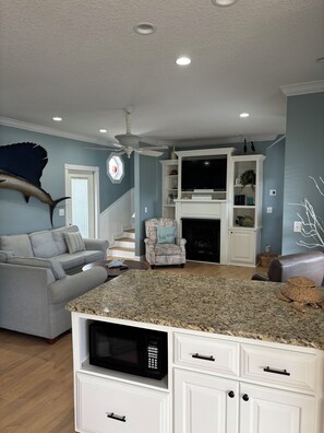 A living room with a grey sofa, a patterned armchair, a kitchen island with a microwave, and a fireplace. - Custom Home on LakeFront in Ocean Lakes (Myrtle Beach)