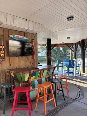 An outdoor seating area with a wooden table and colorful chairs, a wooden wall with a sign, and a view of a house and trees. - Custom Home on LakeFront in Ocean Lakes (Myrtle Beach)