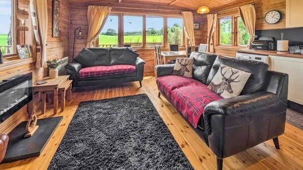 A cozy cabin interior with a fireplace, a sofa with a plaid blanket, a dining area, and large windows offering a view of the outdoors. - 2 bedroom accommodation in Black Isle (Black Isle)