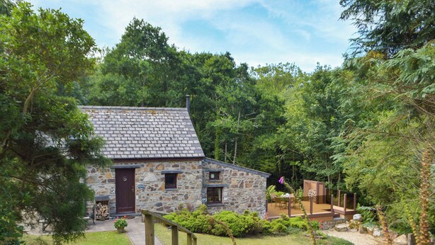 A stone cottage with a sloped roof, surrounded by lush greenery and a well-maintained garden.
