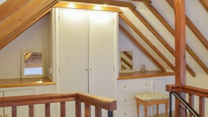 A room with a sloped ceiling, wooden beams, a white wardrobe, a small table with a cushion, and a staircase with wooden railings.
