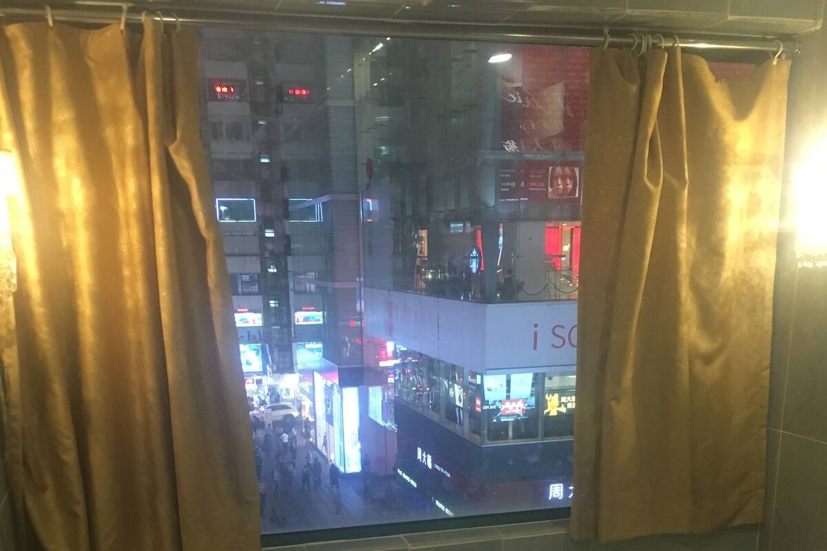 Habitación de hotel con dos camas, una ventana con cortinas, una mesita con una botella y pañuelos, y vistas a un paisaje urbano con edificios iluminados.