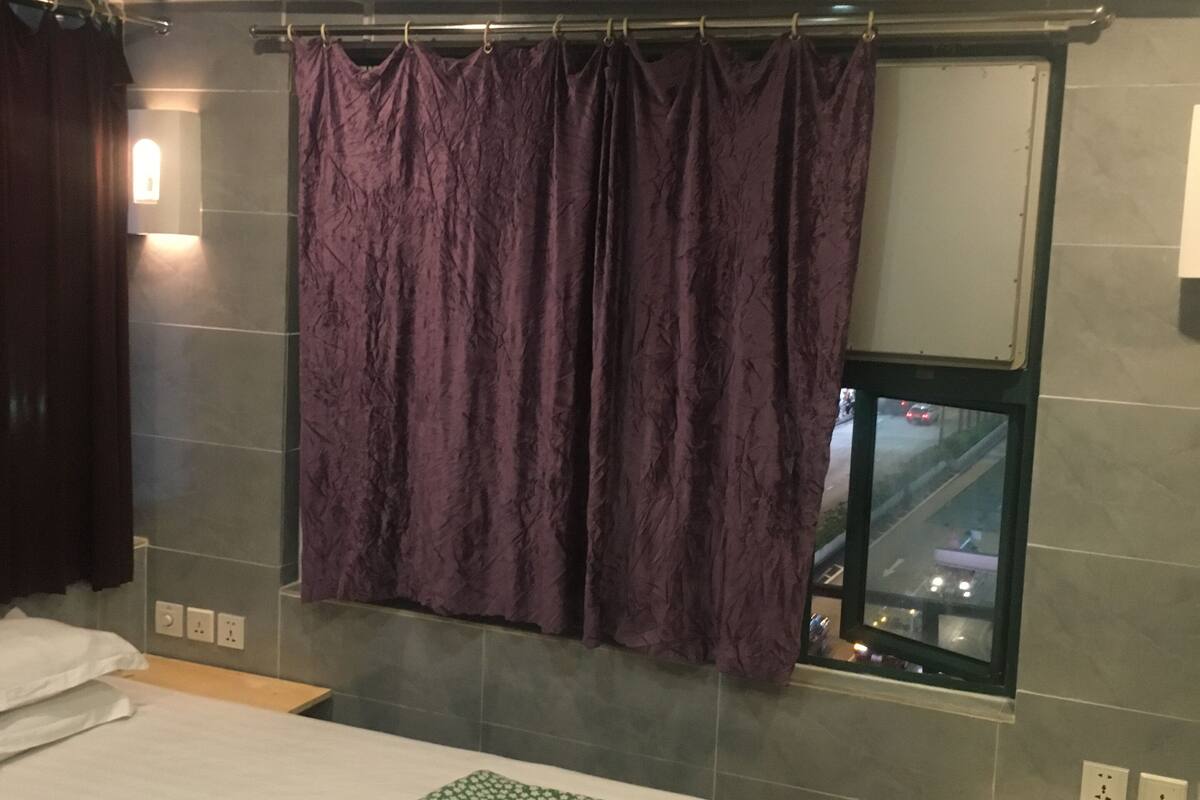 Habitación de hotel con cama, ventana con vistas a una calle concurrida, cortina y un pequeño estante con artículos de aseo.
