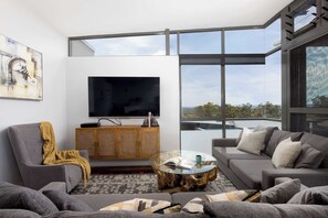 A modern living room with a large flat-screen TV, grey sofas, a glass coffee table, and a wooden cabinet.