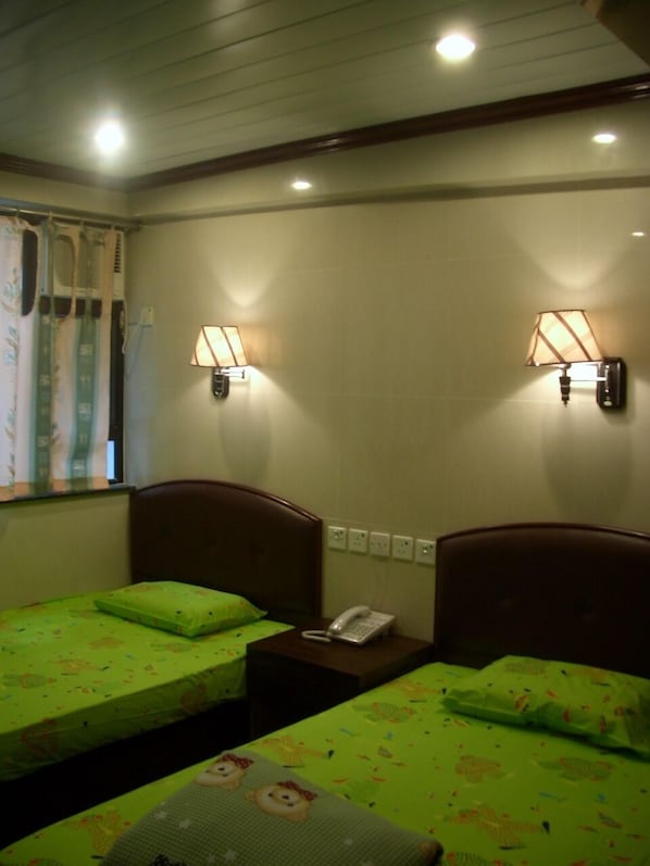 A hotel room with two beds, each with a green bedspread and pillows, a bedside table with a telephone, and wall-mounted lamps.