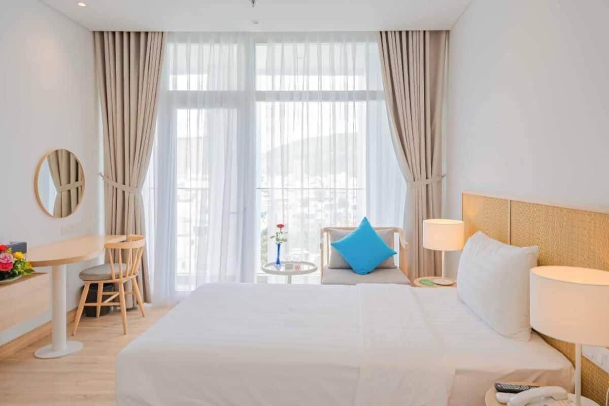 Een hotelkamer met een groot bed, een bureau met stoel, een kleine tafel met bloevasen en uitzicht door het raam.