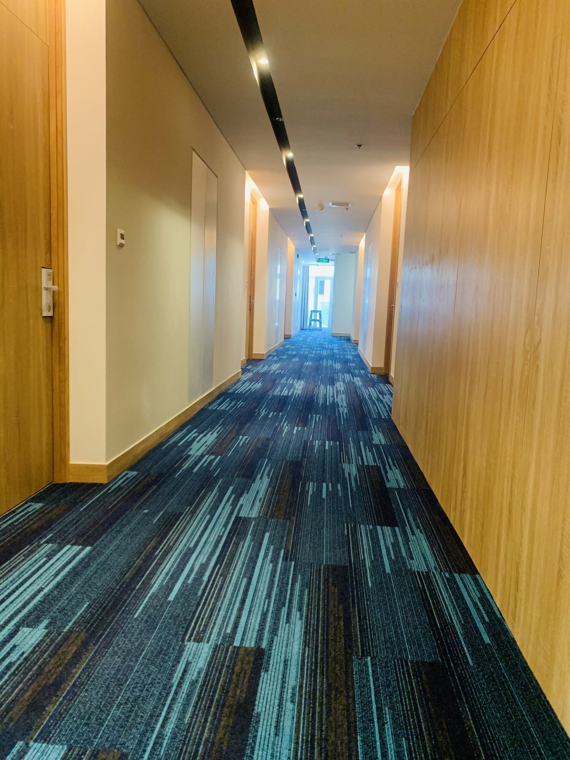 A hotel corridor with a patterned carpet, wooden walls, and a ceiling with recessed lighting.