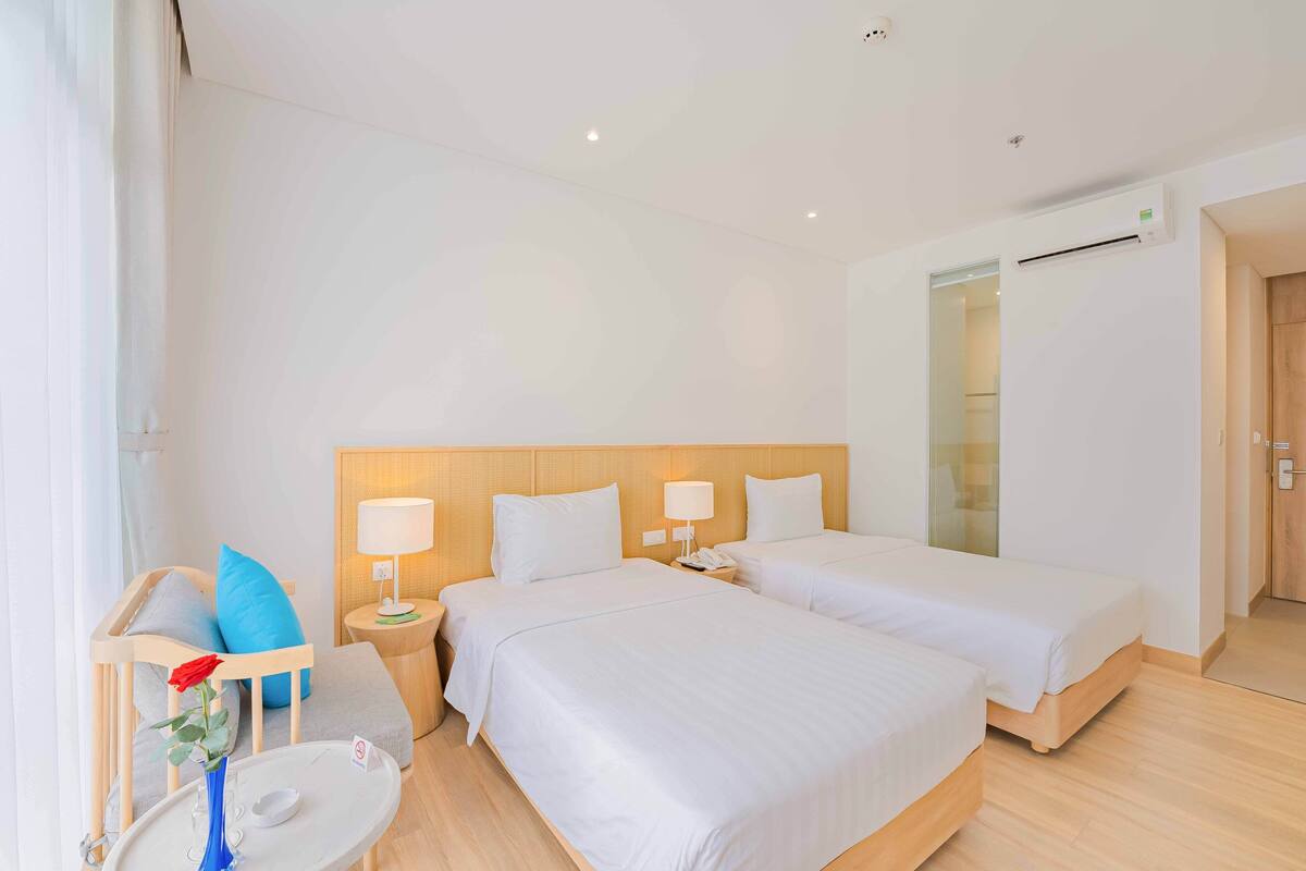 Een hotelkamer met twee bedden, een nachtkastje met een lamp, een tafeltje met een vaas en een telefoon, en een wandairconditioner.