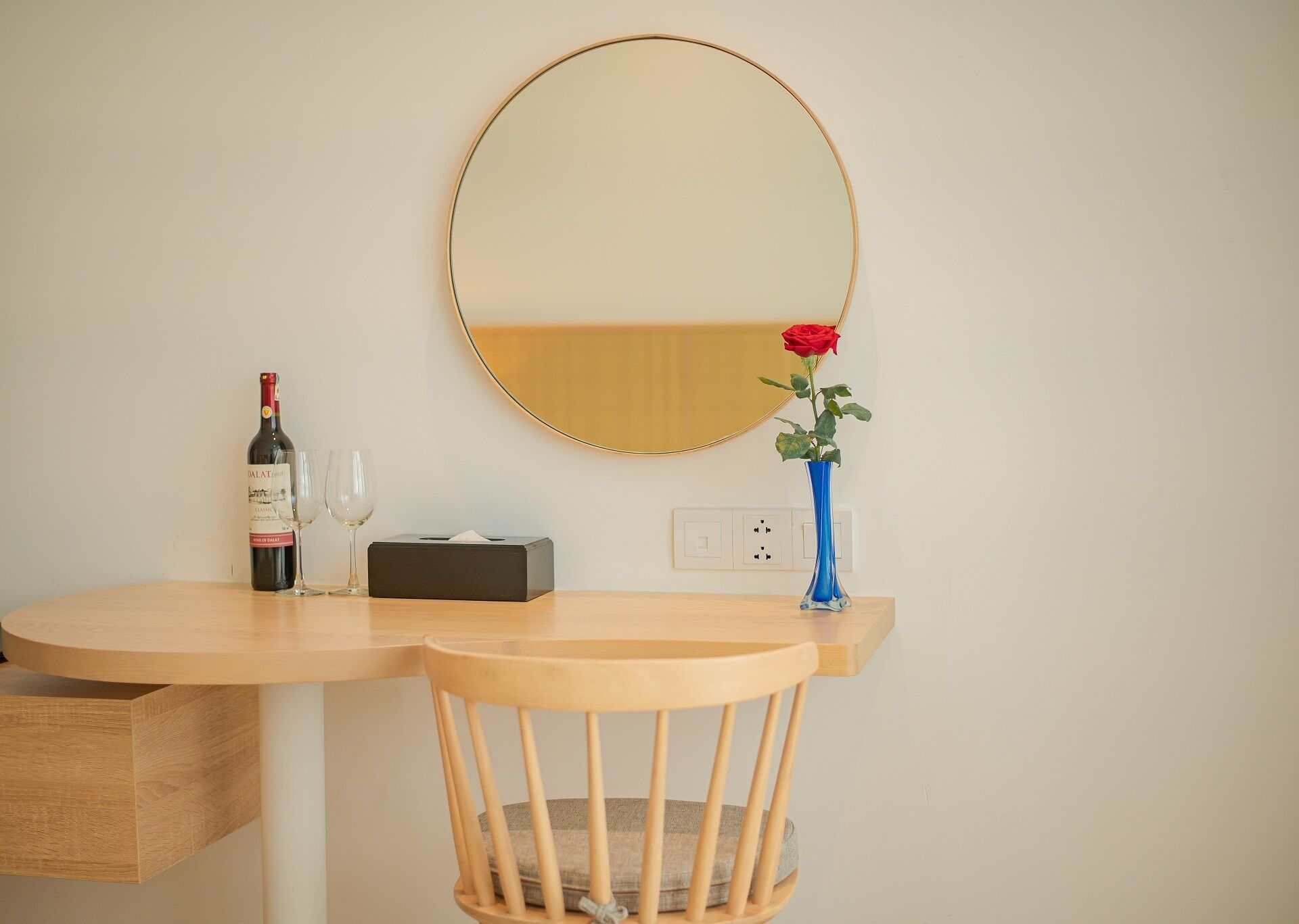 Escritorio de madera con un espejo, una botella de vino, una copa, una caja de pañuelos y un florero con una rosa.