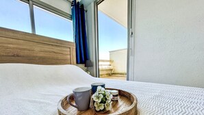 Een net opgemaakt bed met een dienblad vol ontbijt, waaronder een mok, een bord met bloemen en een potje jam, in een kamer met een groot raam en uitzicht op de lucht.