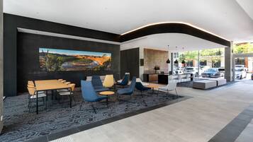 A modern hotel lobby with a large painting, a dining area with chairs and tables, and a view of parked cars outside.
