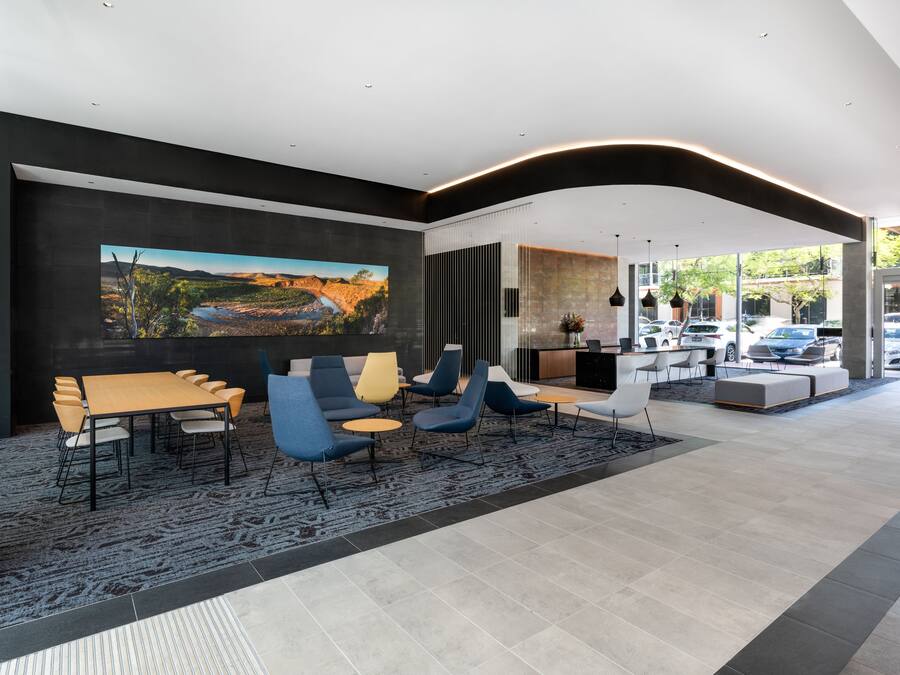 A modern hotel lobby with a large painting, a dining area with chairs and tables, and a view of parked cars outside.