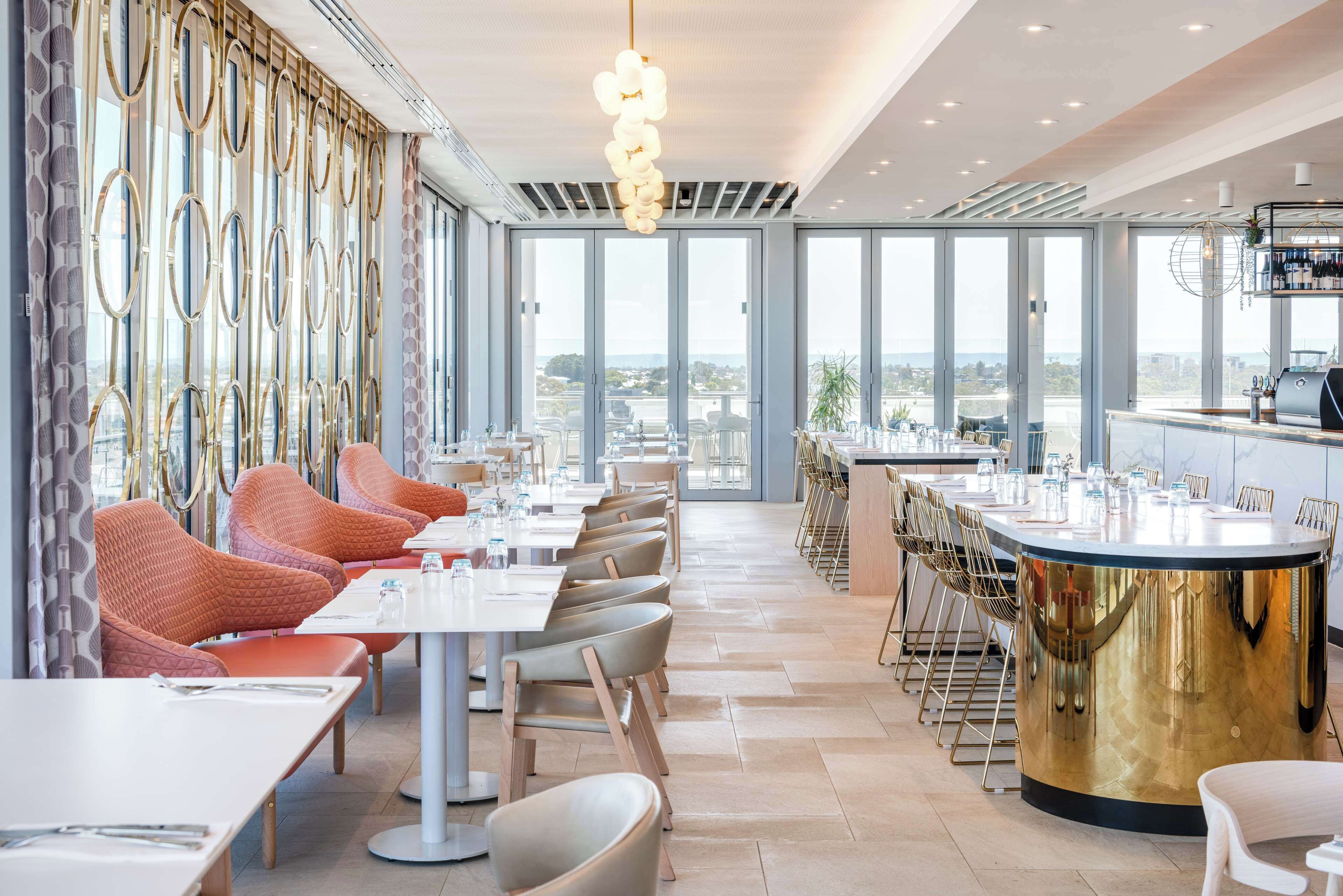 Un restaurante moderno con un amplio comedor, ventanas grandes y una barra con mostrador dorado.