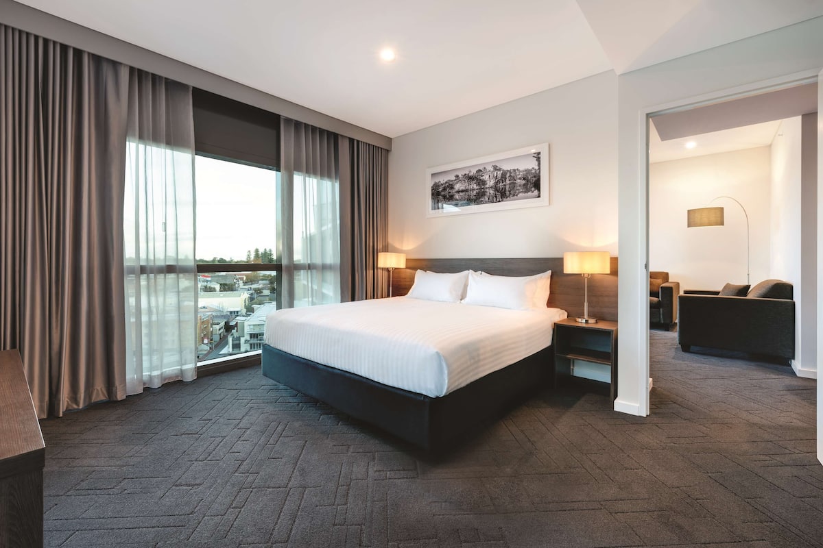 A modern hotel room with a large bed, two bedside tables with lamps, a view of the city through floor-to-ceiling windows, and a framed picture on the wall.