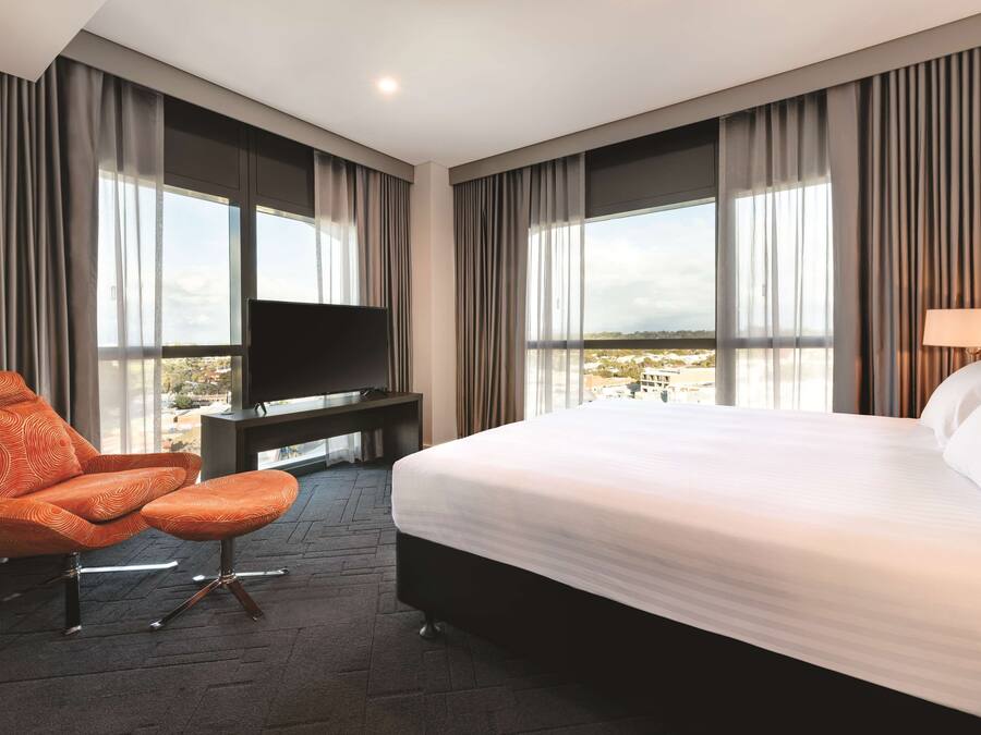 A hotel room with a large bed, a TV on a stand, an orange chair, and a view of the city through large windows.