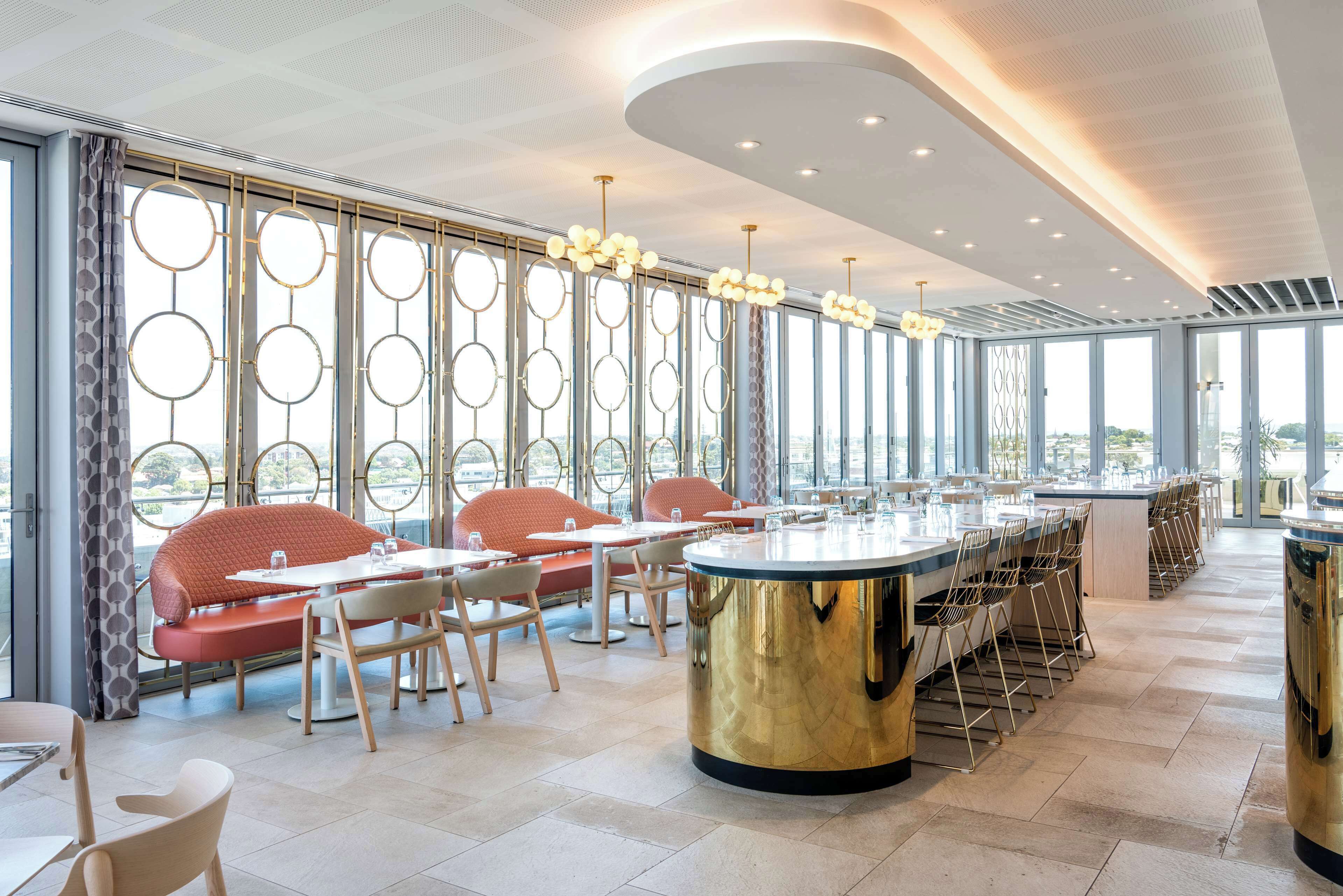 Un restaurante moderno con una barra larga, varias zonas de estar y amplios ventanales con vistas a la ciudad.