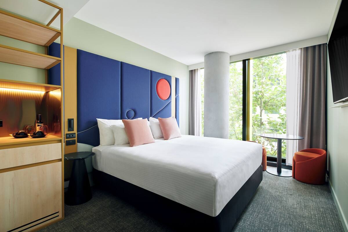 Habitación de hotel con una cama grande, un escritorio con lámpara, una mesita y vistas al exterior con vegetación.