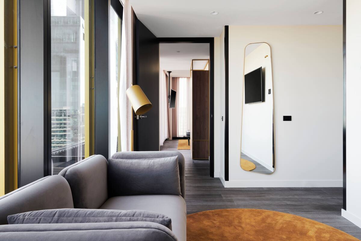 Una habitación de hotel moderna con un sofá, un espejo grande y vistas de la ciudad a través de amplios ventanales.