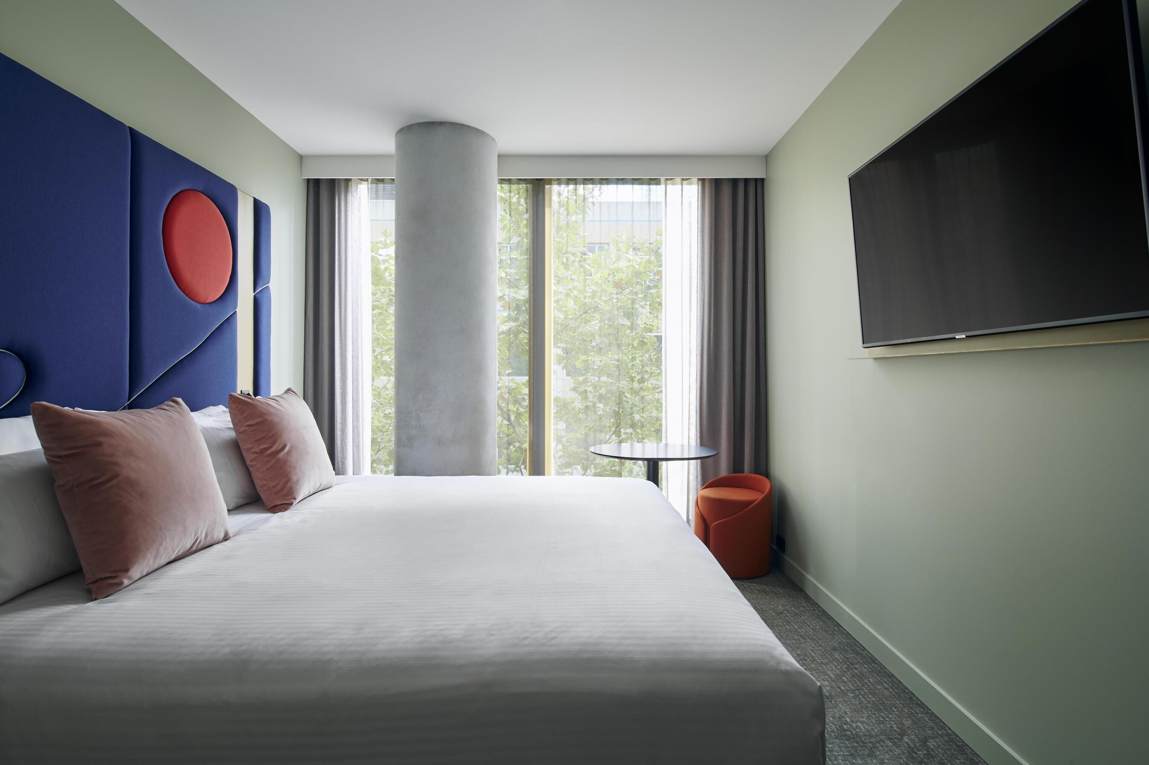 A hotel room with a large bed, a TV, a window with curtains, and a red chair.