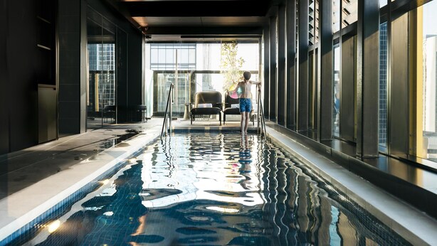 An indoor swimming pool with clear glass walls, a view of the city, and a person standing by the pool. - Quincy Hotel Melbourne (Melbourne)