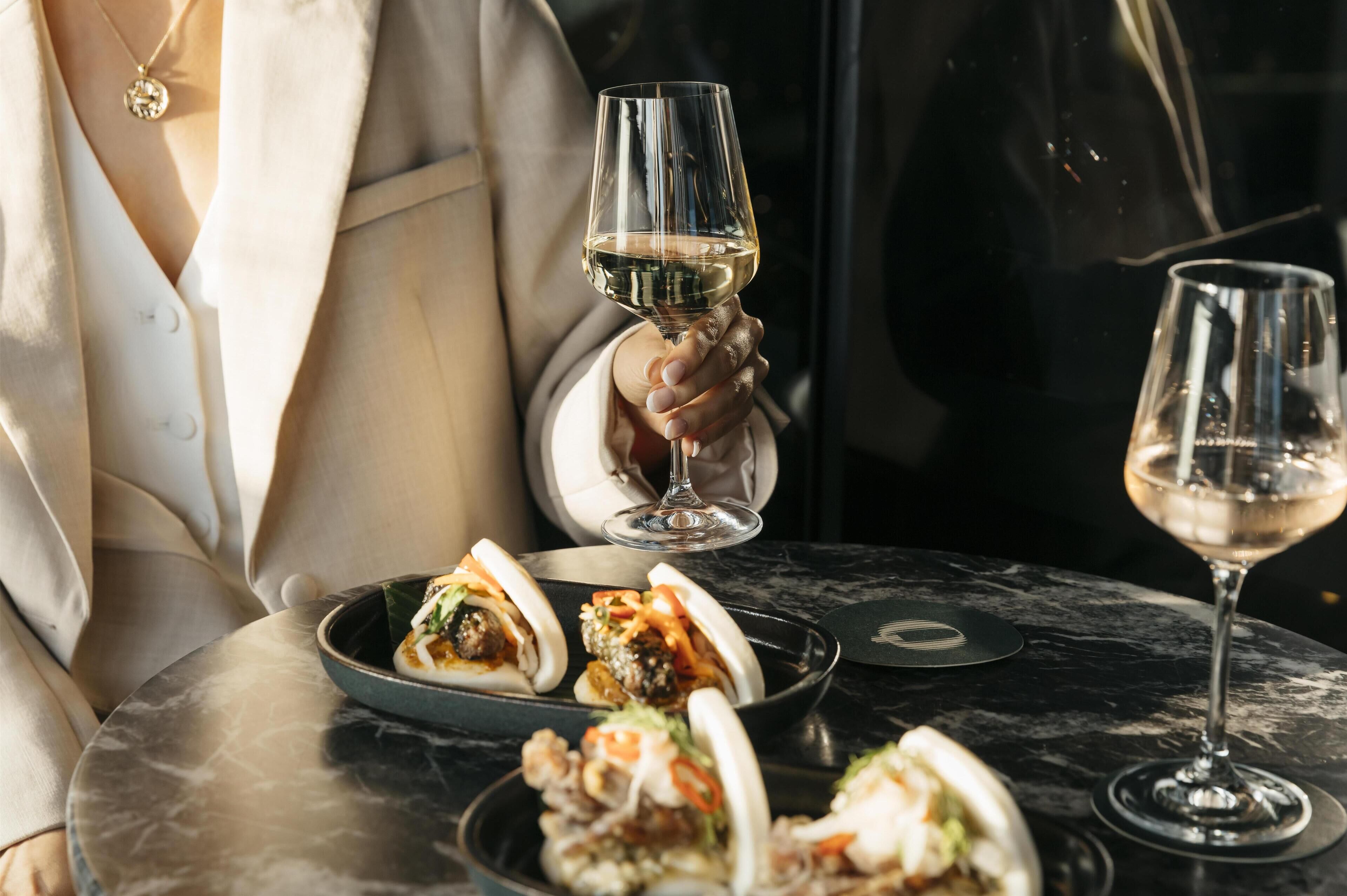 Une personne tenant un verre de vin, assise à une table devant une assiette de nourriture et un autre verre de vin.