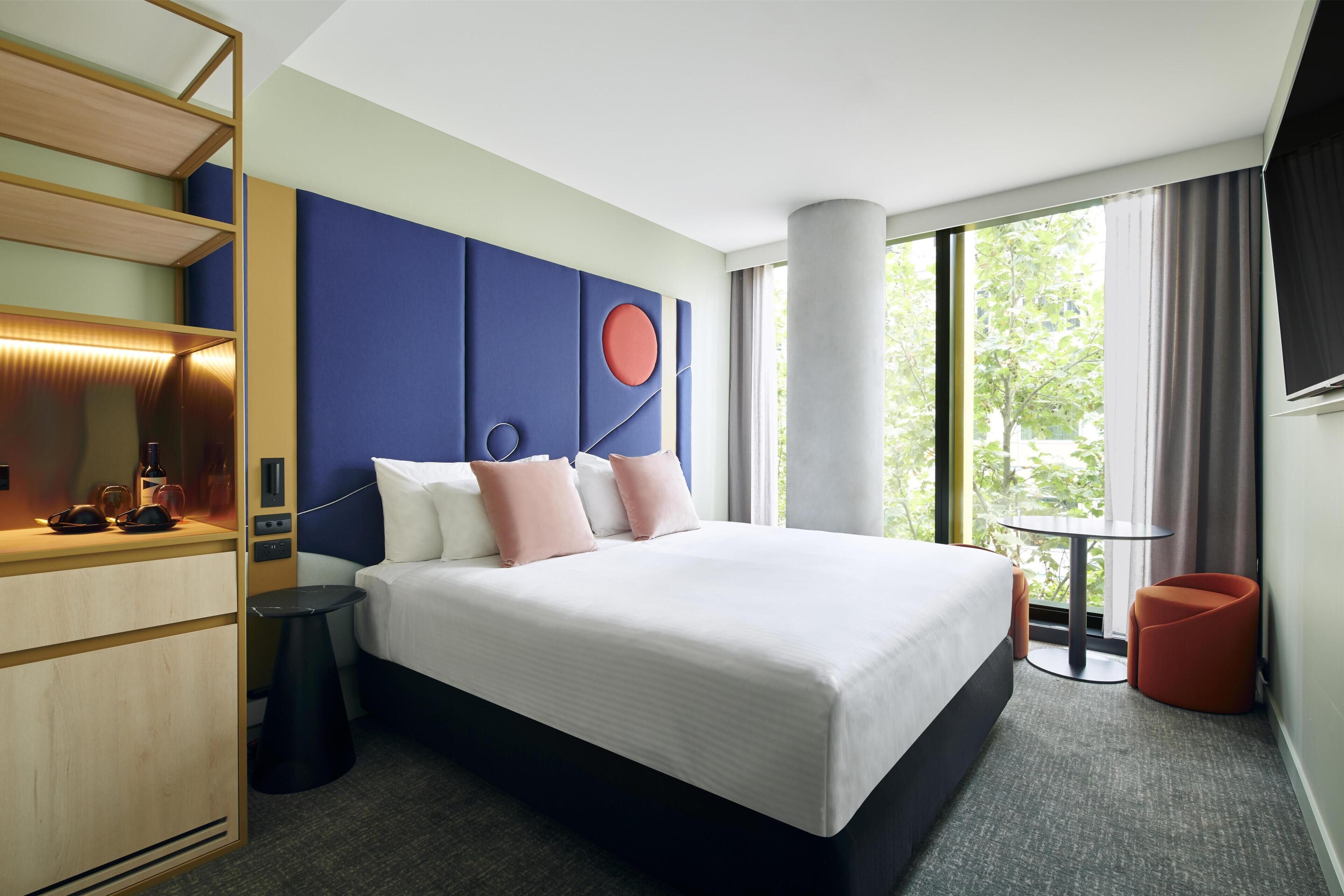 A hotel room with a large bed, a desk with a lamp, a chair, a small table, and a view of greenery through the window.