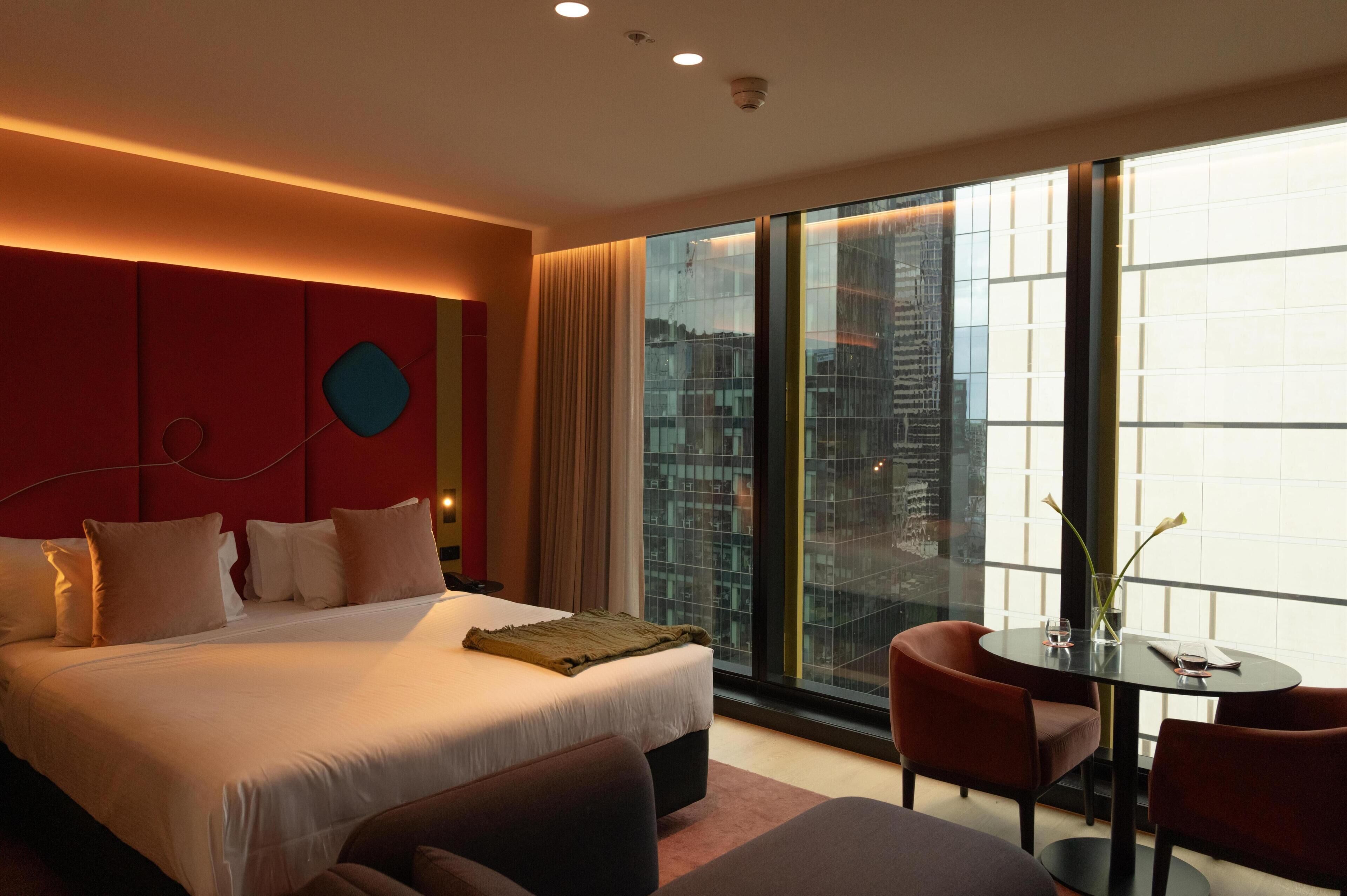 A modern hotel room with a large bed, a small table with chairs, a view of the city through large windows, and a red accent wall.