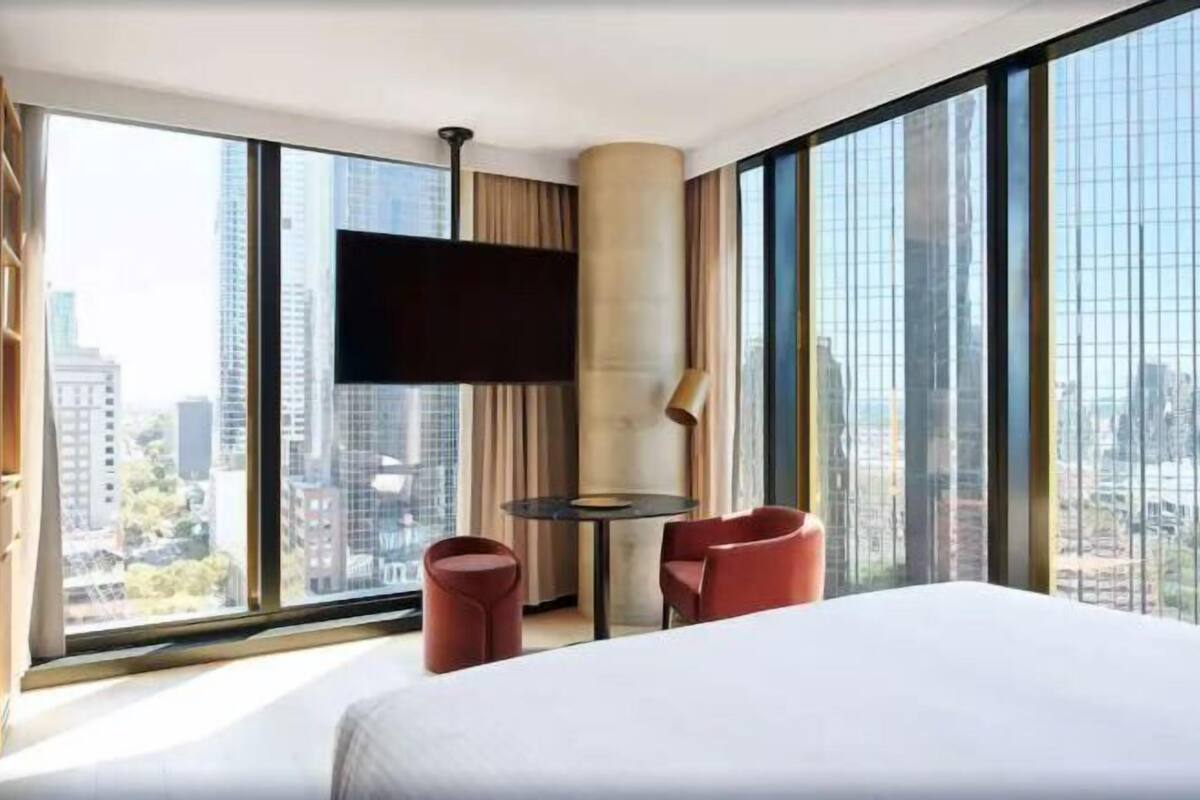 Habitación de hotel con cama, televisor, mesa y sillas, ventanas amplias con vista a la ciudad.