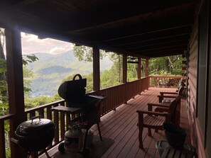 A covered outdoor deck with a grill, chairs, and a table, overlooking a mountainous landscape. - Quaint cabin with privacy and views (Maggie Valley)