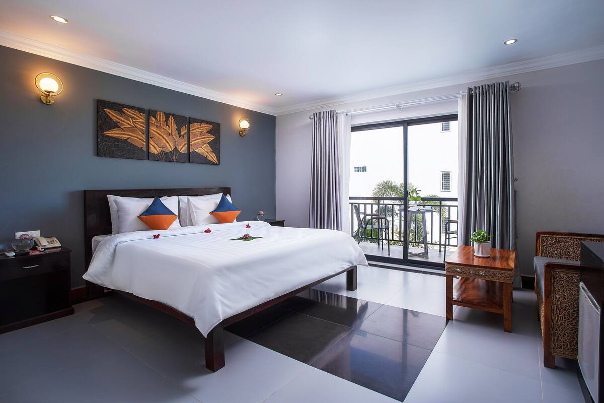 A hotel room with a large bed, a bedside table with a lamp, a small wooden side table, and a view of the outside balcony.
