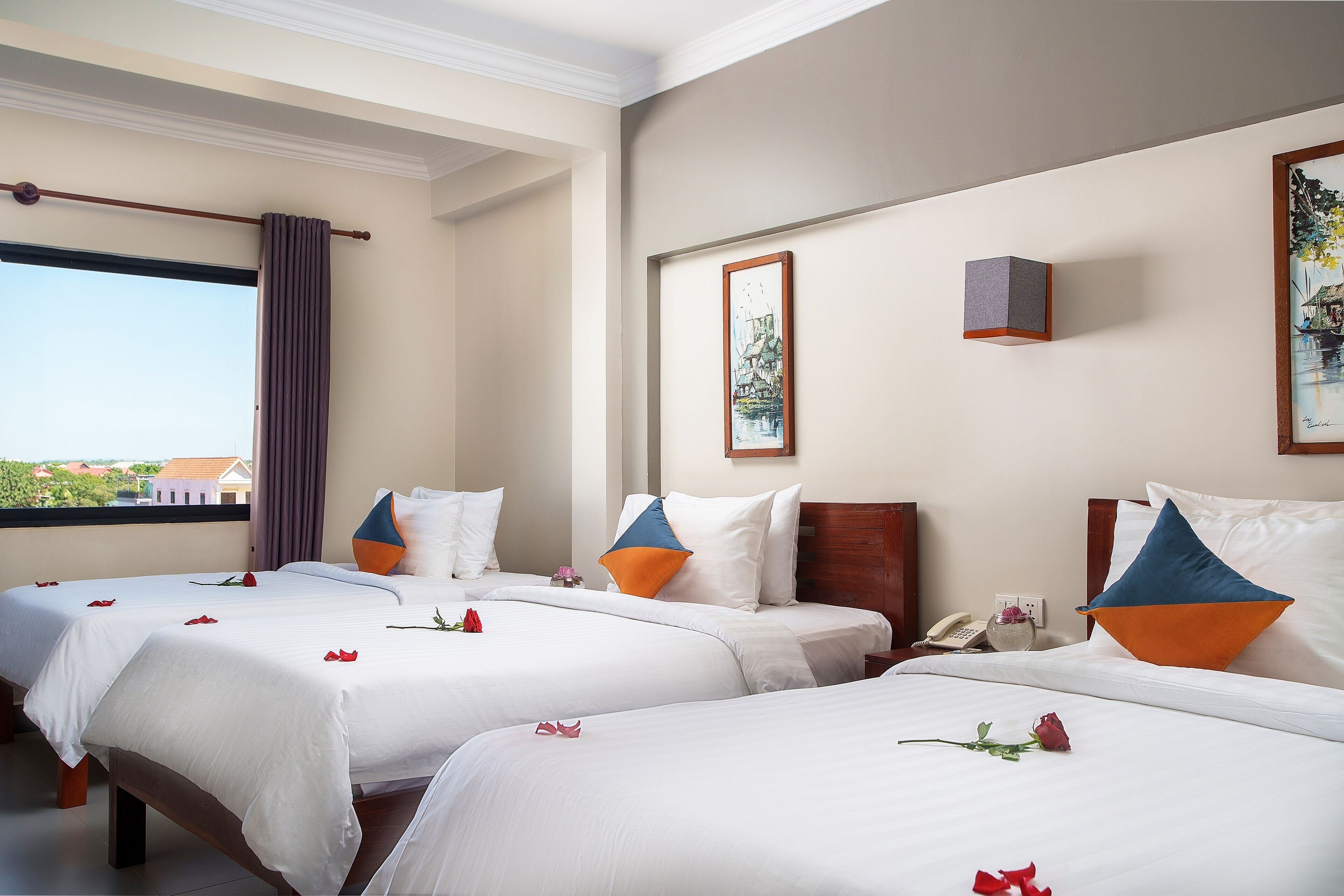 A hotel room with two beds, each with white linens and rose petals. There is a window with a view of buildings and a curtain.