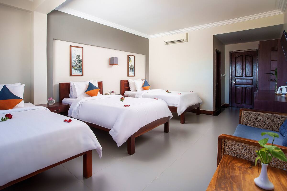 A hotel room with three single beds, a wooden headboard, a small table with a vase, and a door leading to another room.