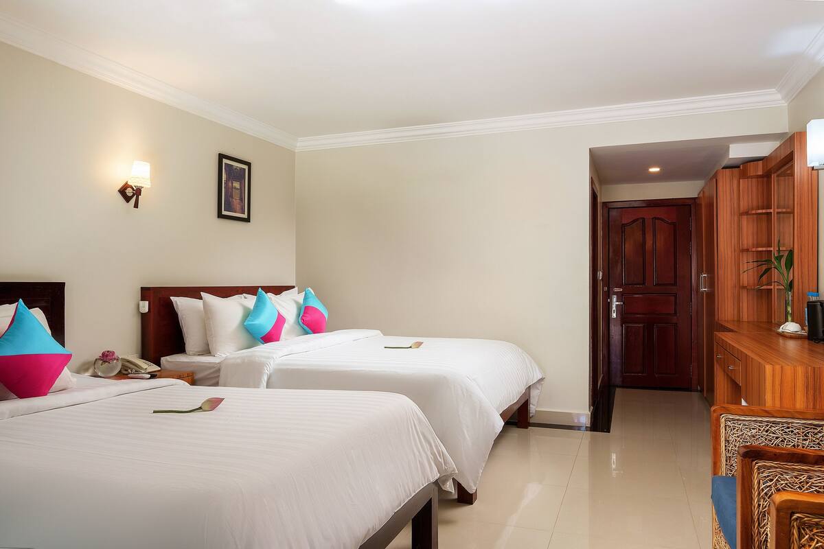 A hotel room with two beds, a wooden desk, a wall-mounted lamp, and a door leading to another room.