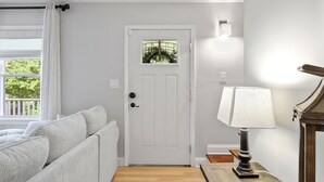 A room with a white door, a window with white curtains, a white sofa, and a table with a lamp.