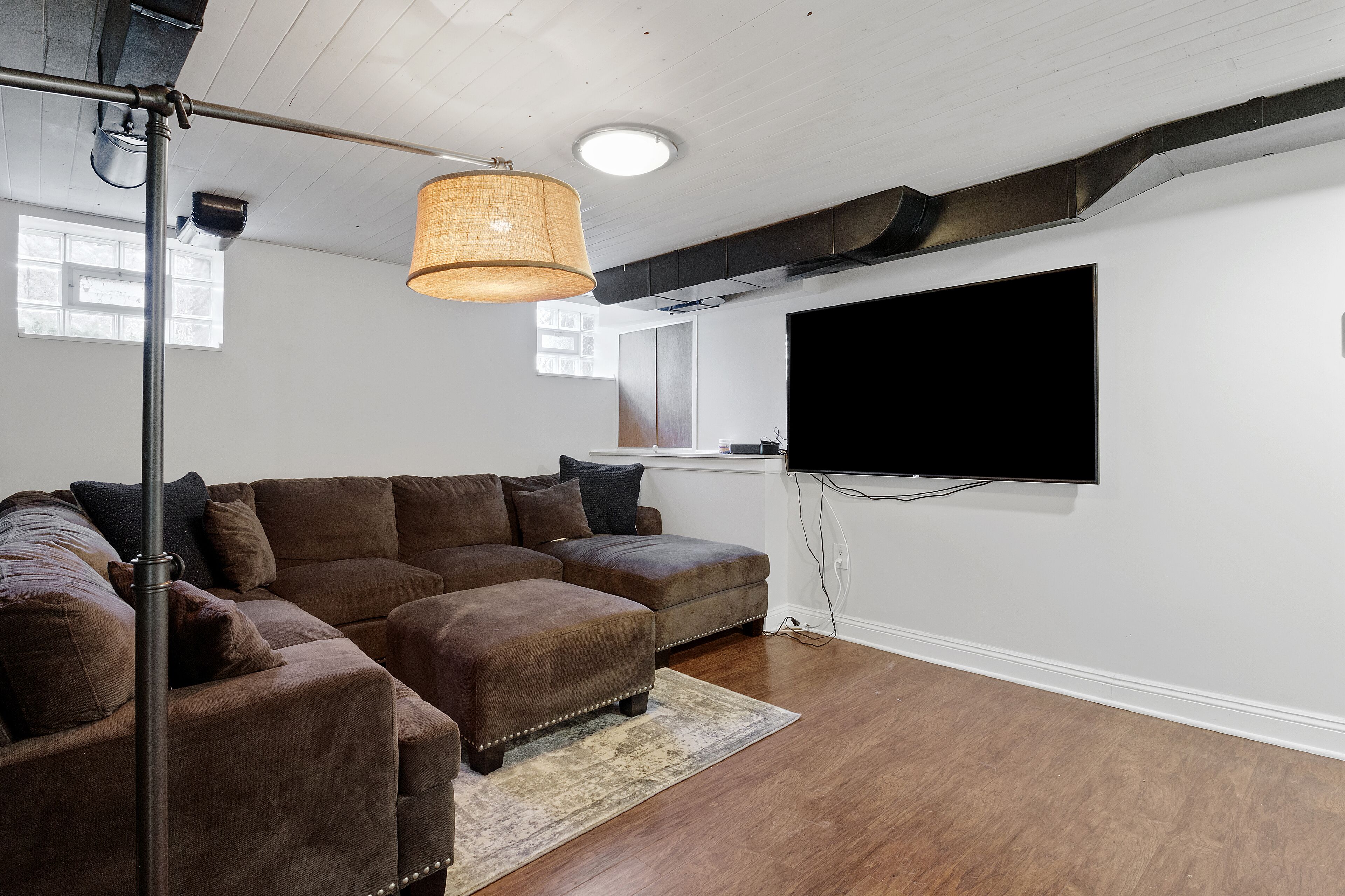 Una sala de estar con un sofá seccional, un televisor de pantalla plana montado en la pared y una lámpara de techo.