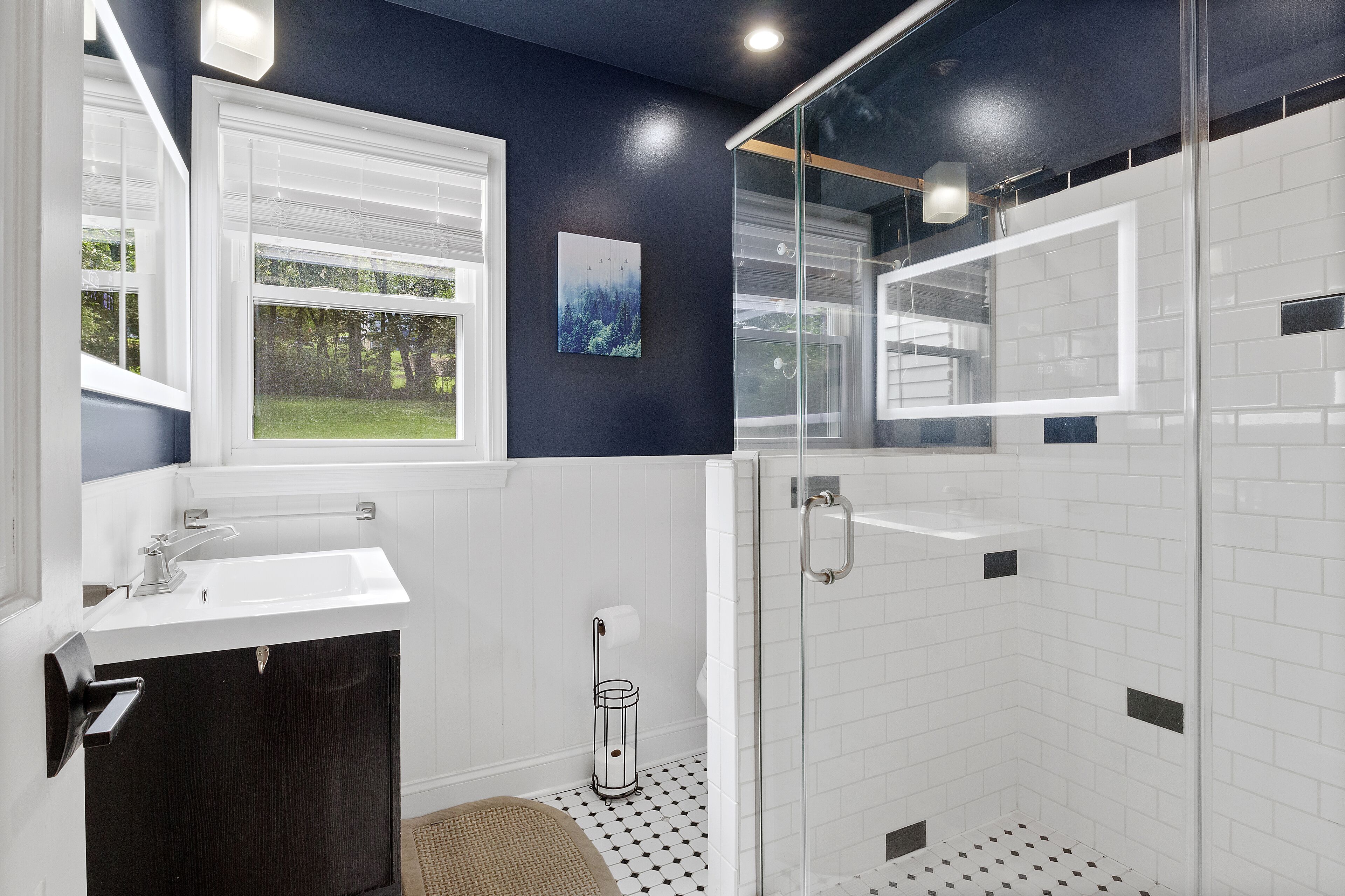 A modern bathroom with a glass shower enclosure, white tiles, a dark blue accent wall, and a window with a view of greenery.