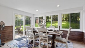 A dining area with a table set for four, surrounded by white chairs with floral cushions, large windows offering a view of a green garden, and a ceiling fan on the left.