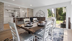 A dining area with a wooden table set for four, surrounded by white chairs with patterned cushions. A brick wall and white cabinetry are visible in the background.