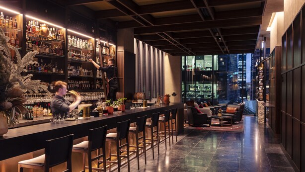 A bar area with a bartender, shelves of bottles, and a seating area with a city view. - Next Hotel Melbourne, Curio Collection by Hilton (Melbourne)