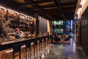 A bar area with a bartender, shelves of bottles, and a seating area with a city view. - Next Hotel Melbourne, Curio Collection by Hilton (Melbourne)