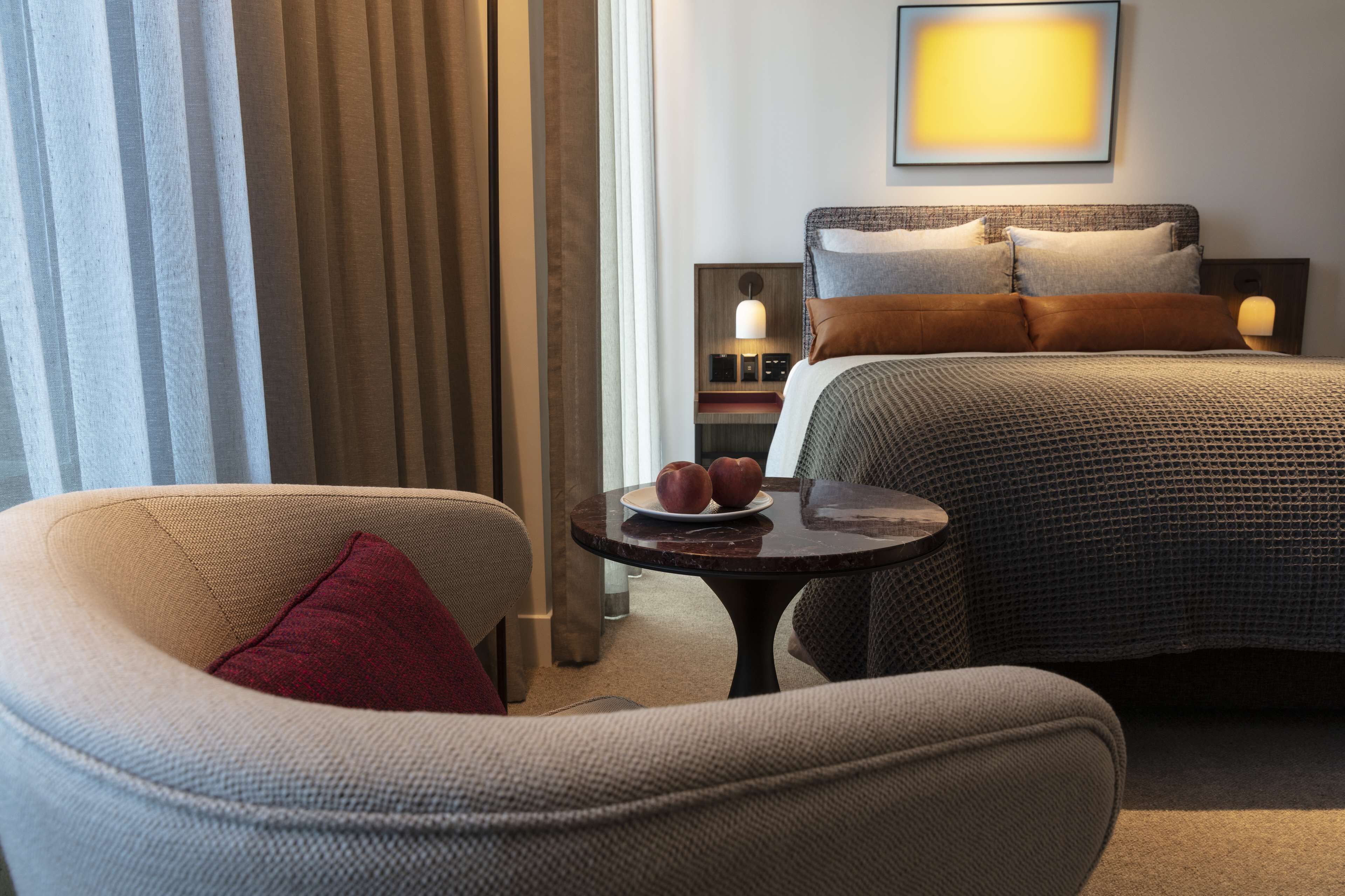 A hotel room with a bed, a chair, a small table, and a bedside table with a lamp.
