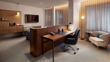 A modern hotel room with a wooden desk, a sofa, a television, and a large window with curtains.